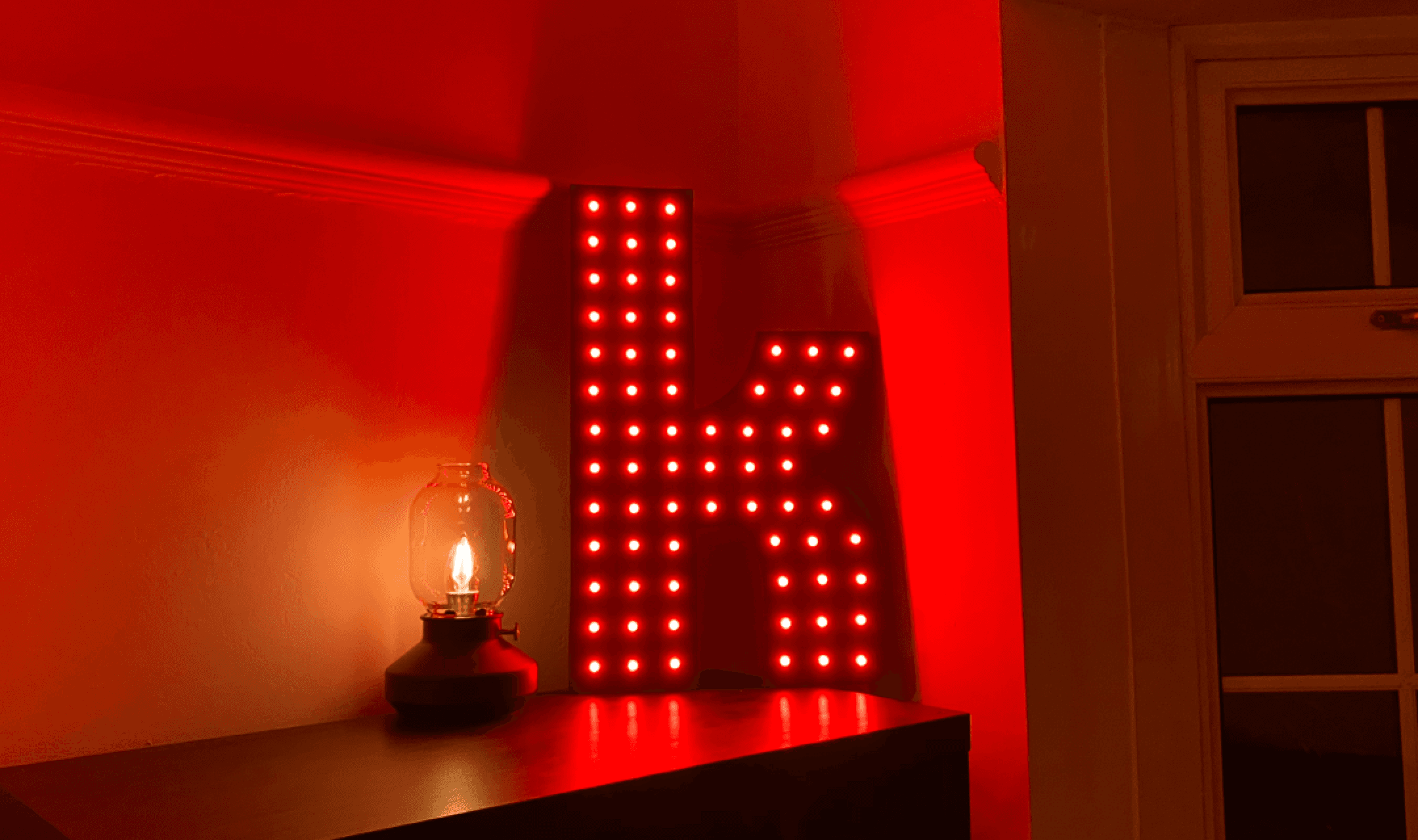 Finished light-up 'K' sign glowing red on a sideboard beside a small lantern lamp